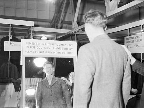 Aircrew training cadet views his new demob suit in a standing mirror with a placard above it saying "Remember in future you may have to use coupons - choose wisely. Please do not hurry in your choice" at a civilian clothing centre in Wembley, 17 October, 1944